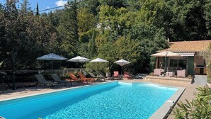 Ein Swimmingpool mit Liegestühlen und Sonnenschirmen, umgeben von viel Grün und einem Gebäude mit einem Ziegeldach.