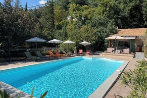 En swimmingpool med solstolar och parasoller, omgiven av grönska och en byggnad med ett tak täckt av tegelpannor.