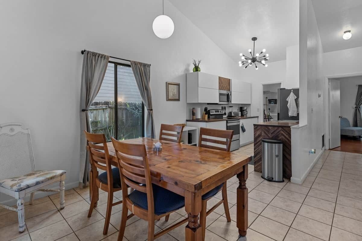 A dining area with a wooden table and chairs, a white chair, a window with curtains, a kitchen area with cabinets and appliances, and a trash bin.