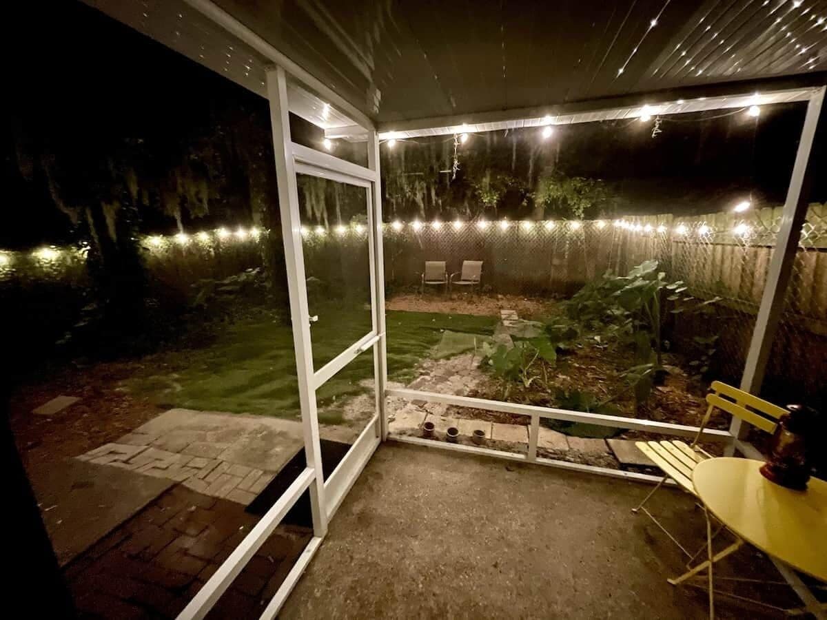 Un espacio al aire libre cubierto con una mesa y sillas, un pequeño jardín y luces colgantes.