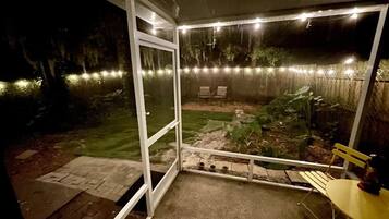 Un espacio al aire libre cubierto con una mesa y sillas, un pequeño jardín y luces colgantes.