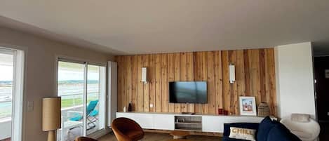 A modern living room with a flat-screen TV, wooden panel wall, and a view of the outdoors.