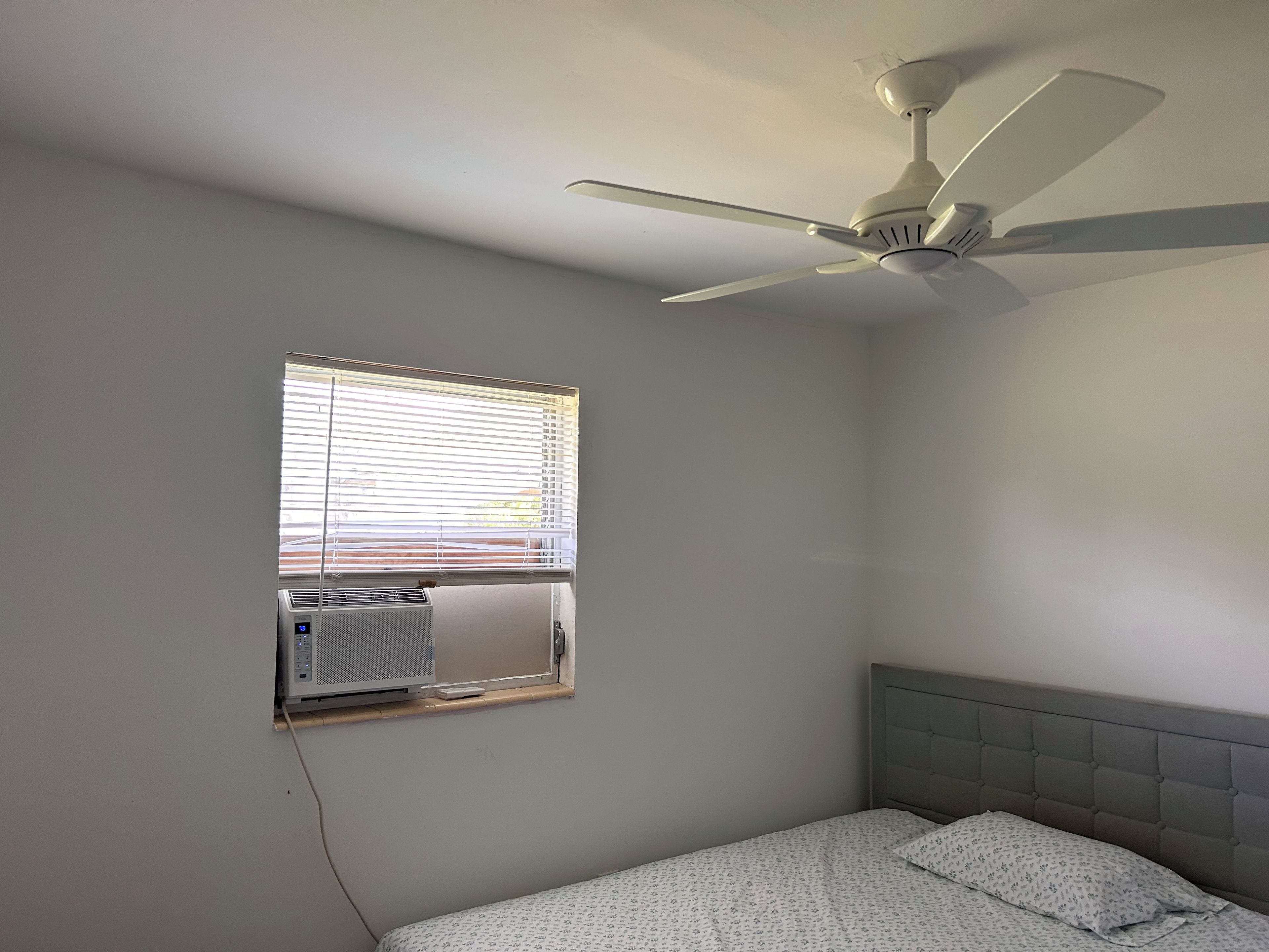 Ein Schlafzimmer mit einem Einzelbett, Deckenventilator, Klimaanlage und einem Fenster mit Jalousien.