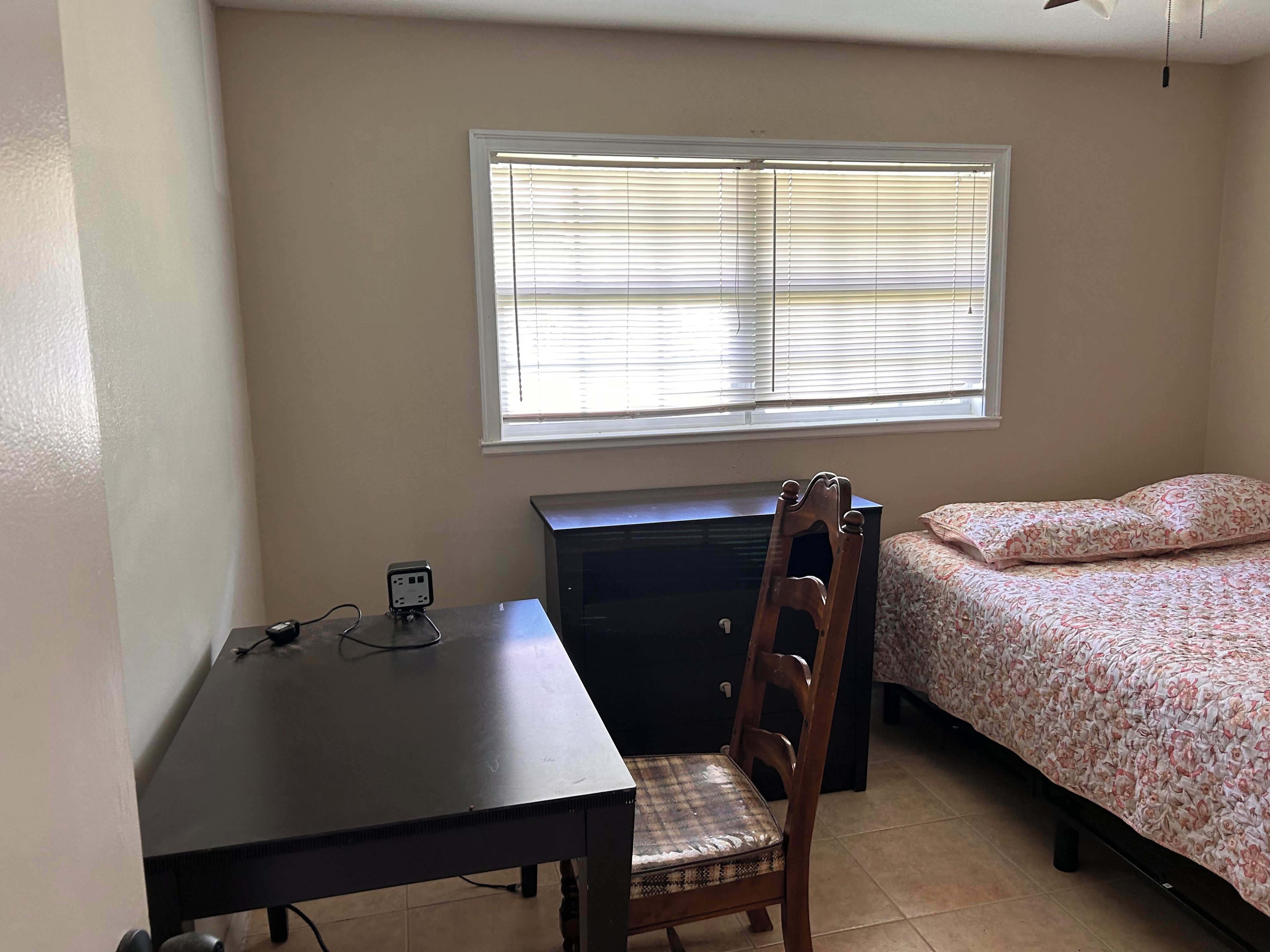 A small room with a bed, a desk, and a chair. There is a window with blinds, a power outlet on the desk, and a ceiling fan.