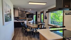 A dining area with a wooden table and chairs, a kitchen in the background, and a view of greenery through large windows. - Mississippi River view. Duck Hunters Get away. 4 Bedroom, 5 bed - Sleeps 10. (Brownsville)