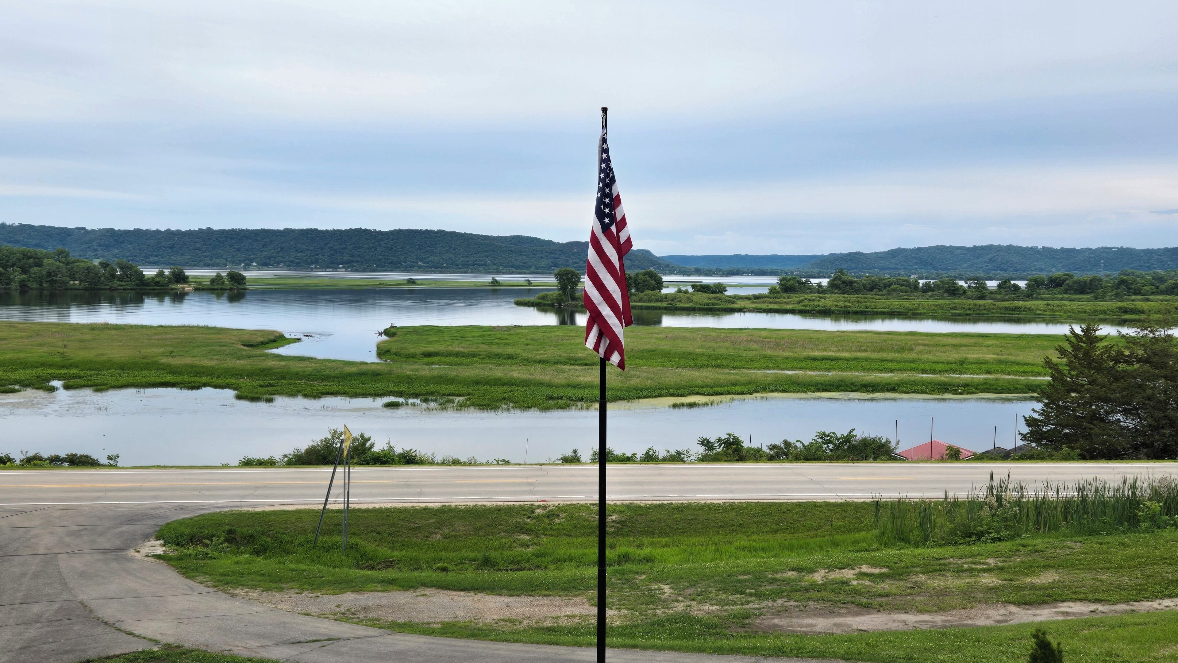 A flagpole with an American flag in a rural setting with a road and water bodies in the background.