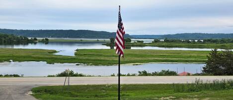 A flagpole with an American flag in a rural setting with a road and water bodies in the background.