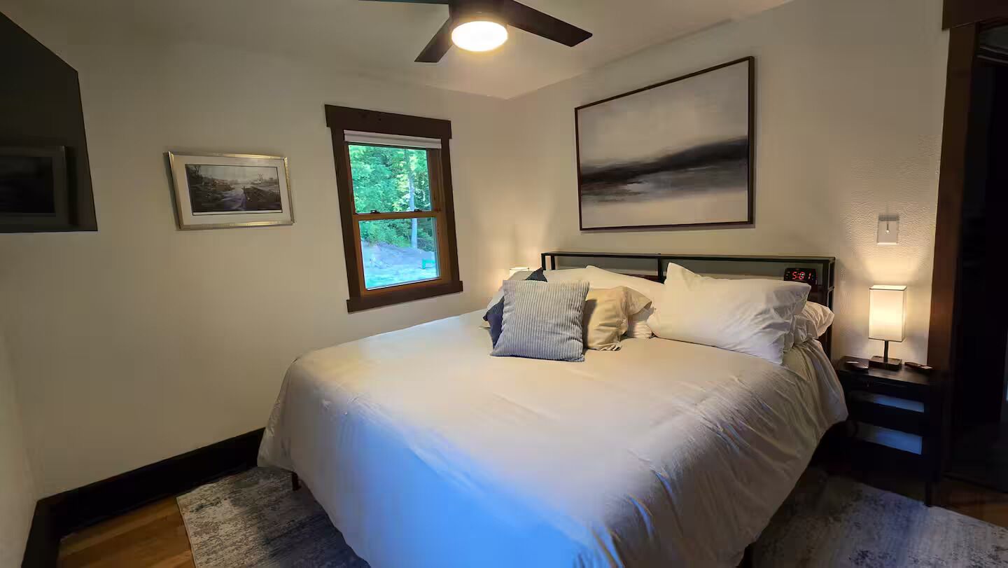 A bedroom with a bed, a window, a ceiling fan, and a nightstand with a lamp.