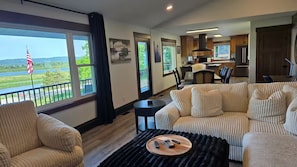 A living room with a sofa, a coffee table with remotes, a view of a river, and a kitchen in the background.