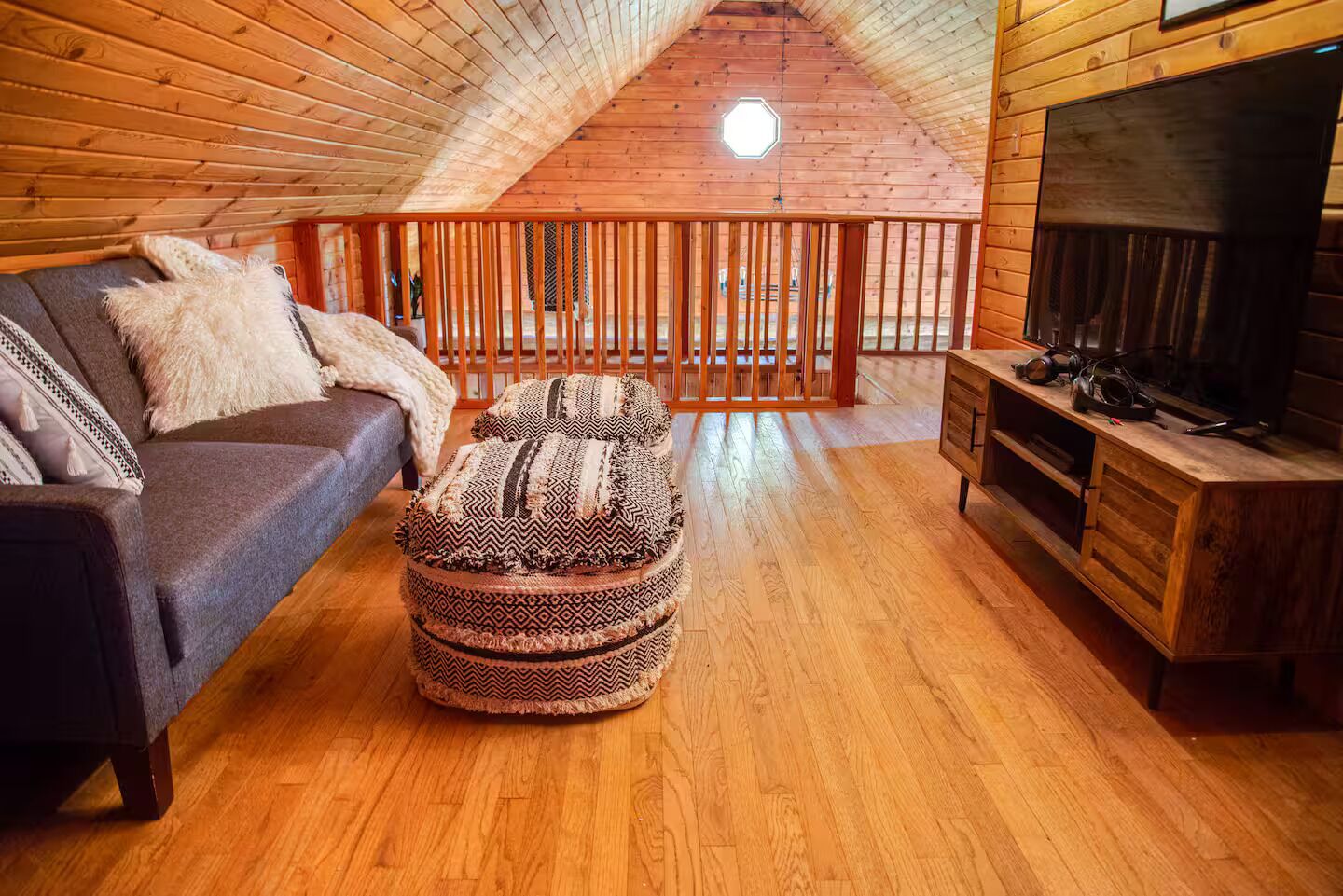 Une pièce mansardée chaleureuse avec un plafond en bois, un canapé, un pouf à motifs, une télévision et une armoire en bois.