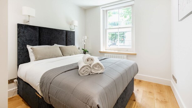 A modern bedroom with a dark headboard, a large window, and a neatly made bed with rolled towels. - Chic 1BR Fitzrovia Apartment by Bloomsbury (London)