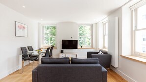 A modern living room with a sofa, a dining table with chairs, a flat-screen TV, and large windows. - Chic 1BR Fitzrovia Apartment by Bloomsbury (London)