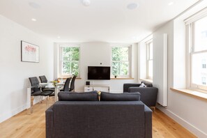 Ein modernes Wohnzimmer mit einer Couch, einem Esstisch mit Stühlen, einem Flachbildfernseher und großen Fenstern.