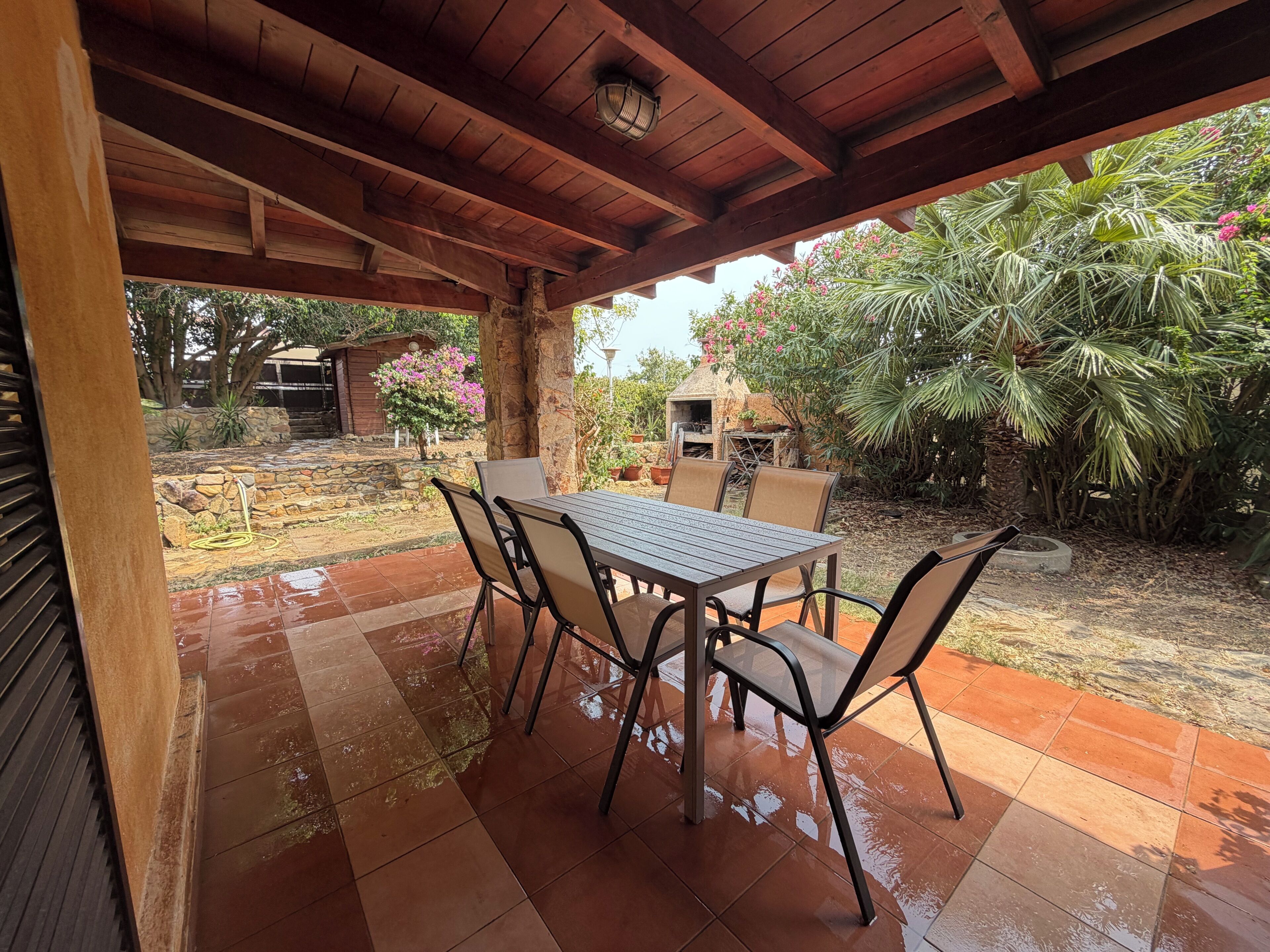Une terrasse extérieure avec une table et des chaises, entourée d’un jardin où poussent différentes plantes et où se trouve une petite construction.