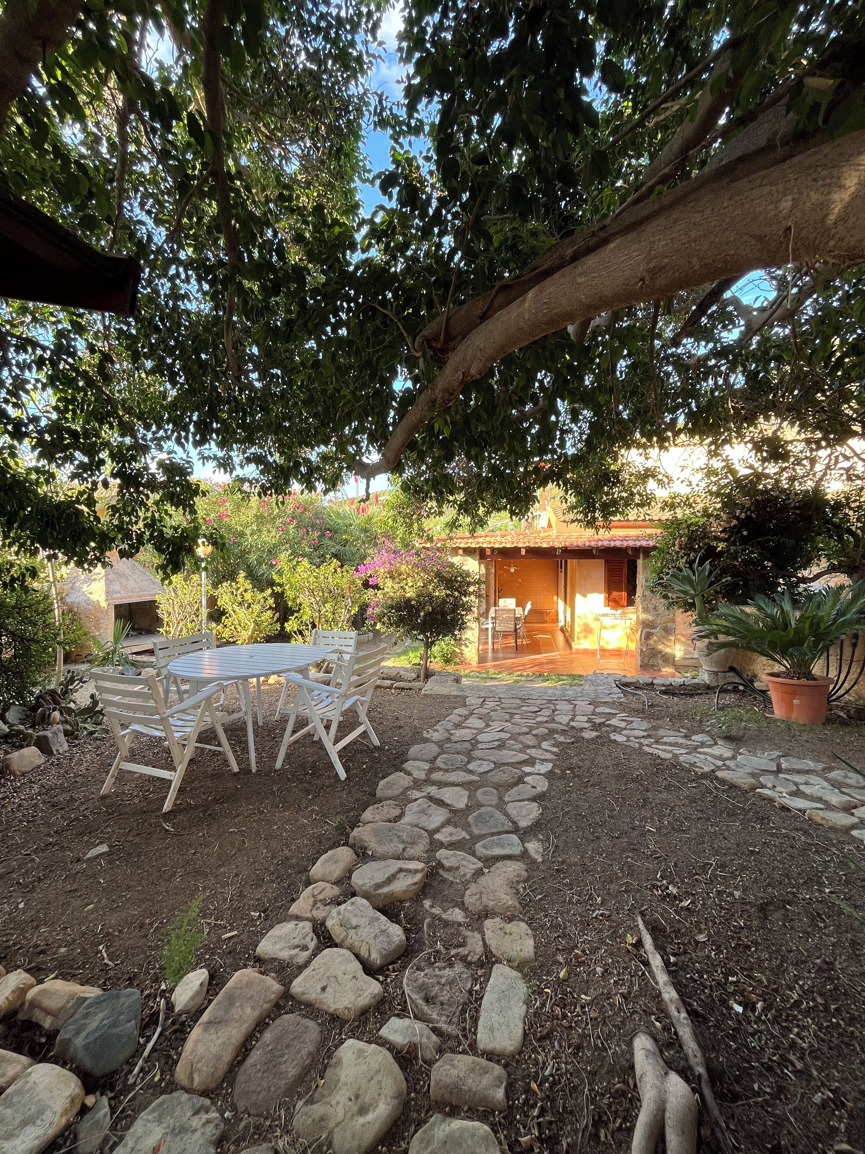 A stone pathway leading to a wooden table and chairs set under a tree, with a building in the background.
