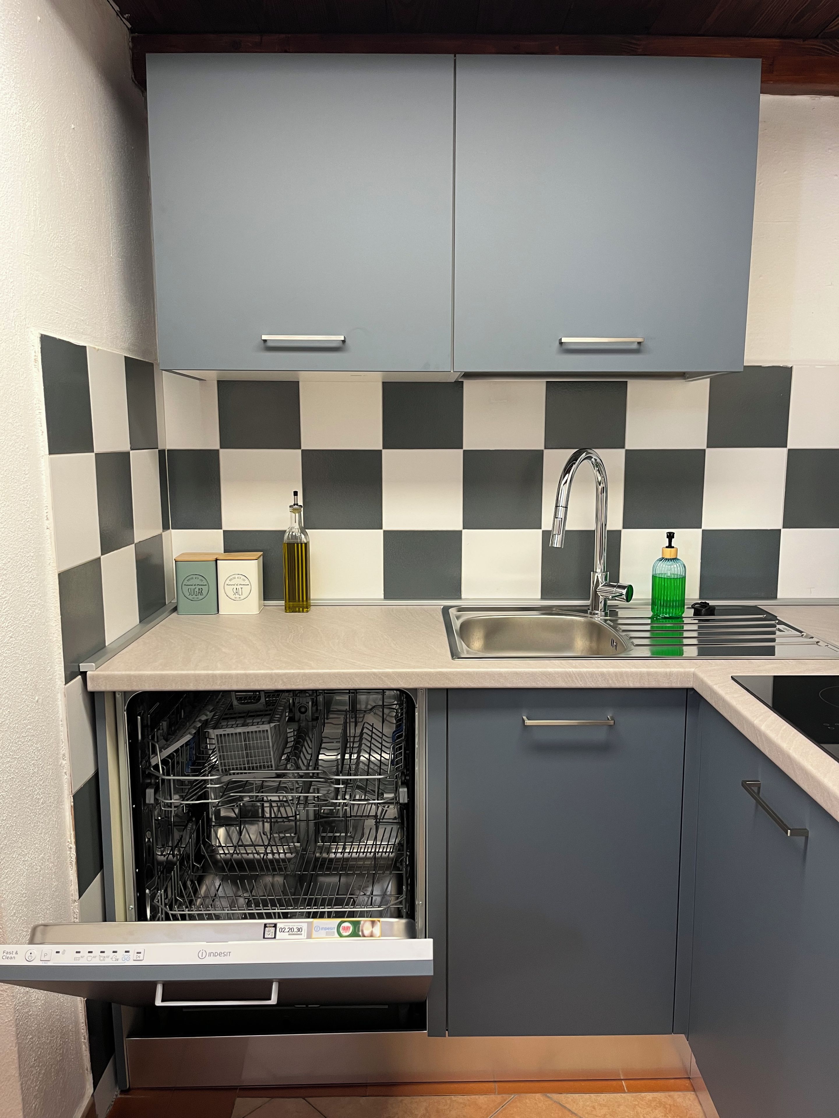 Une cuisine compacte avec des meubles gris, un dosseret à carreaux, un évier en acier inoxydable et un lave-vaisselle.