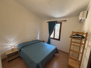 A small room with a bed, a nightstand, a window with curtains, a wooden ladder, and a wall-mounted air conditioner. - Villa Aurea House with Garden, Vico Andrea Doria Bassa 8 (Costa Rei)