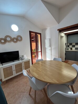 A dining area with a round wooden table, white chairs, a television on a cabinet, and a kitchen visible through an open door. - Villa Aurea House with Garden, Vico Andrea Doria Bassa 8 (Costa Rei)