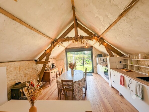 An attic room with a dining table, chairs, a kitchen area with cabinets and appliances, and a view through a large window.