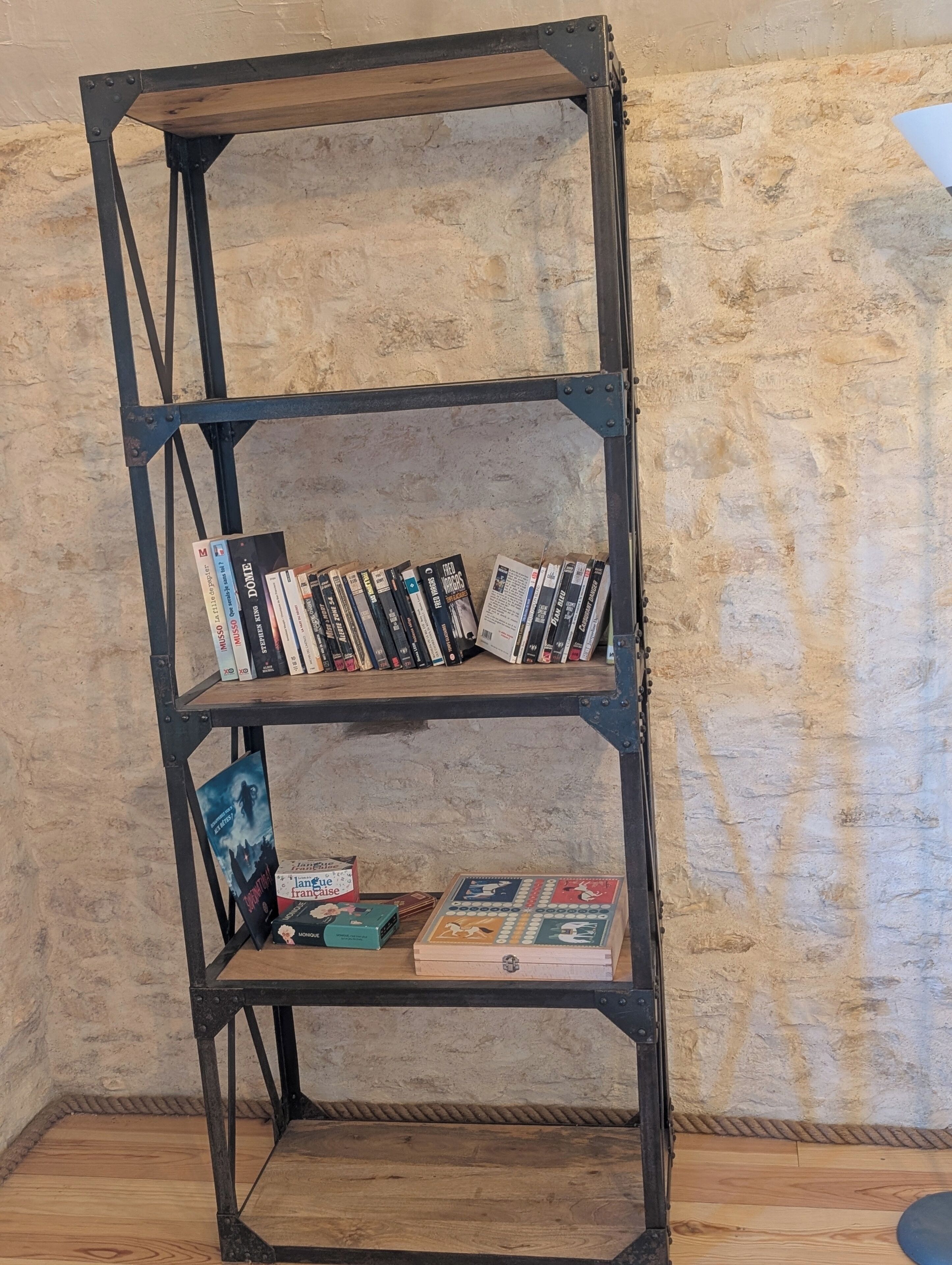 A metal bookshelf with books and a board game against a textured wall.