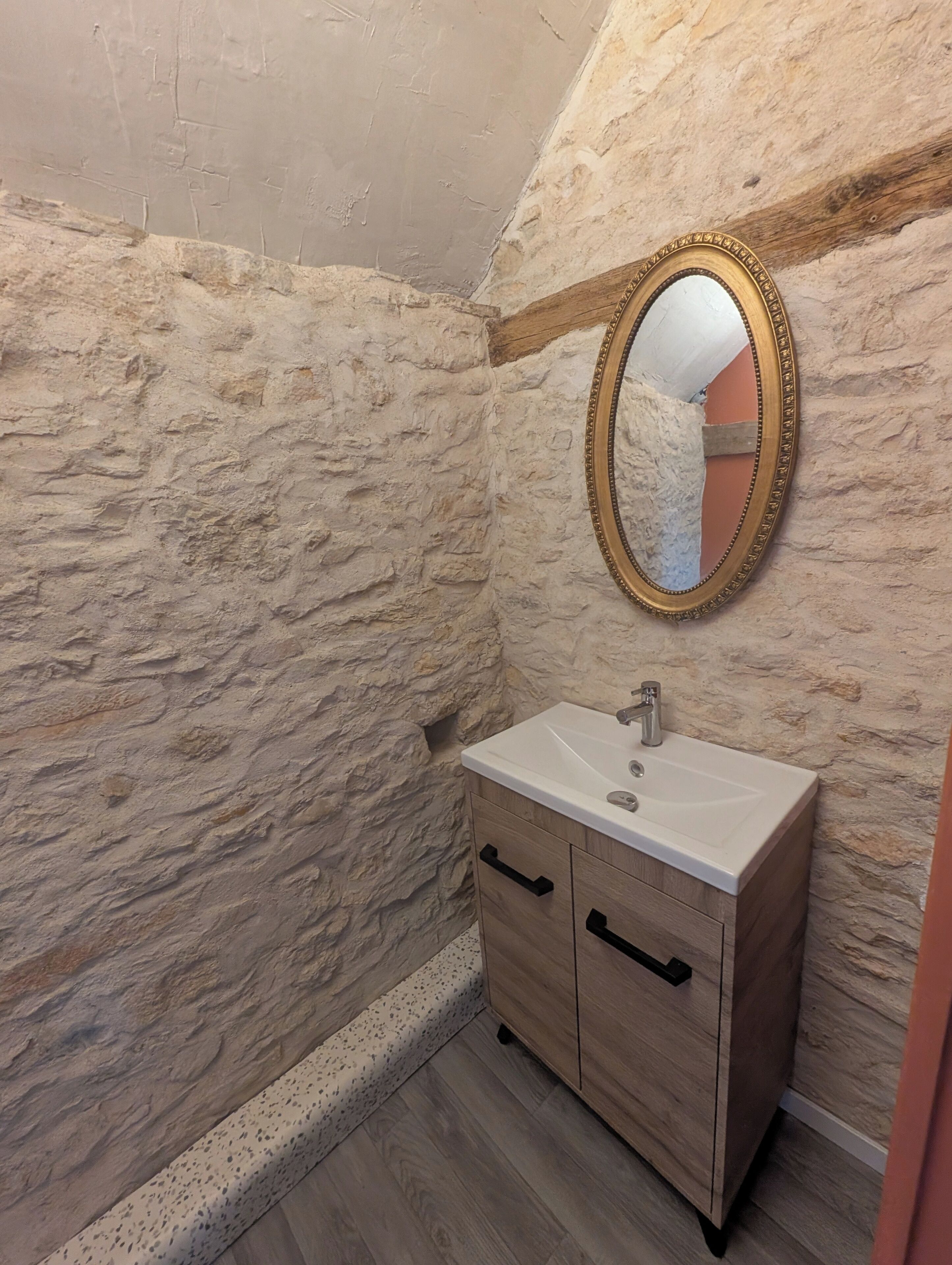 Un bagno con parete in pietra, mobiletto in legno con lavello e specchio con cornice dorata.