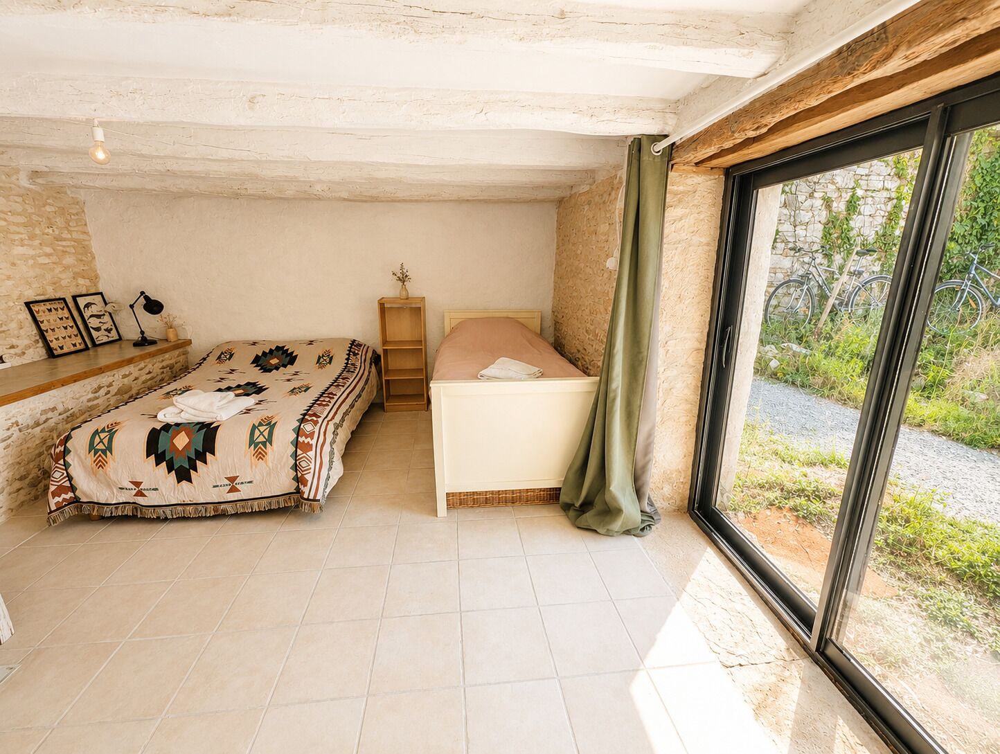 A bedroom with a bed, a bedside table, a lamp, and a window with a view of greenery.
