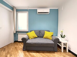 A room with a grey sofa, two yellow cushions, a white radiator, a window, and a small white table with a plant. - Principino - Walk to the Old Port and the Aquarium; take the train to the Cinque Terre. (Genova)