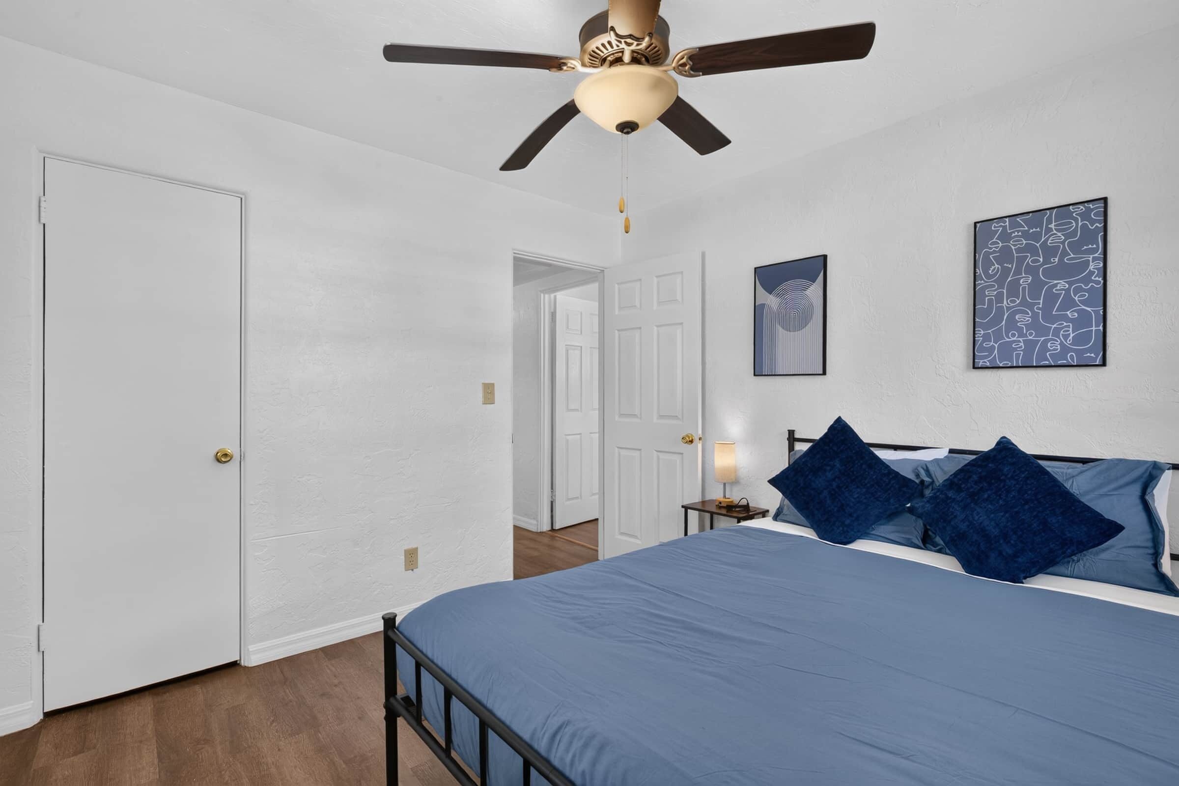 A bedroom with a bed, a ceiling fan, a door, and a wall with two framed pictures.