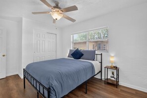 A bedroom with a bed, a ceiling fan, a window with blinds, and a bedside table with a lamp.