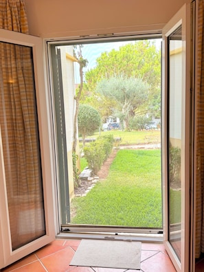 A sliding glass door leading to a grassy outdoor area with a view of a tree and parked cars. - Fantastic Apartment: " Azul " in Tavira, with a garden terrace and swimming pool (Tavira)