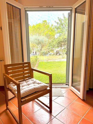 Une chaise en bois avec un coussin, dans une pièce dont le sol est recouvert de carreaux, et qui offre une vue sur un espace extérieur grâce à une porte-fenêtre coulissante, donnant sur une pelouse et des arbres.