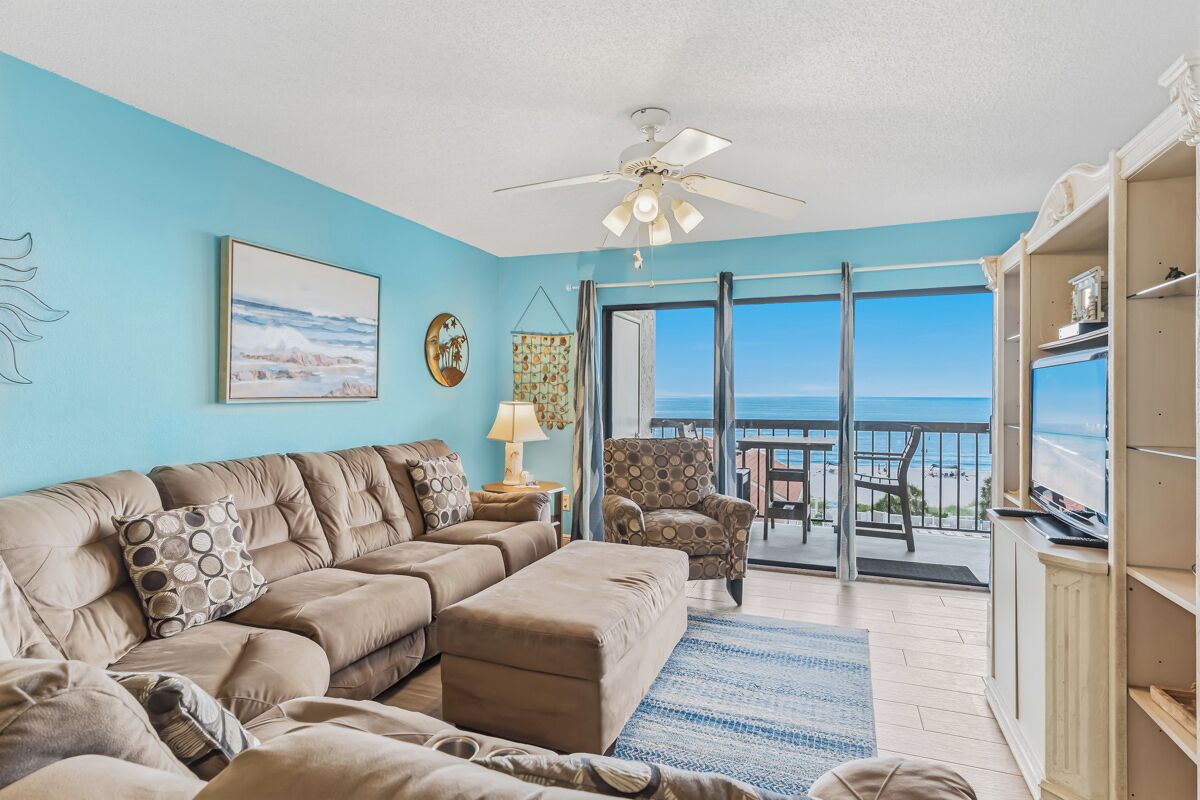 A living room with a sectional sofa, a ceiling fan, a view of the ocean through a sliding glass door, and a balcony with outdoor furniture.