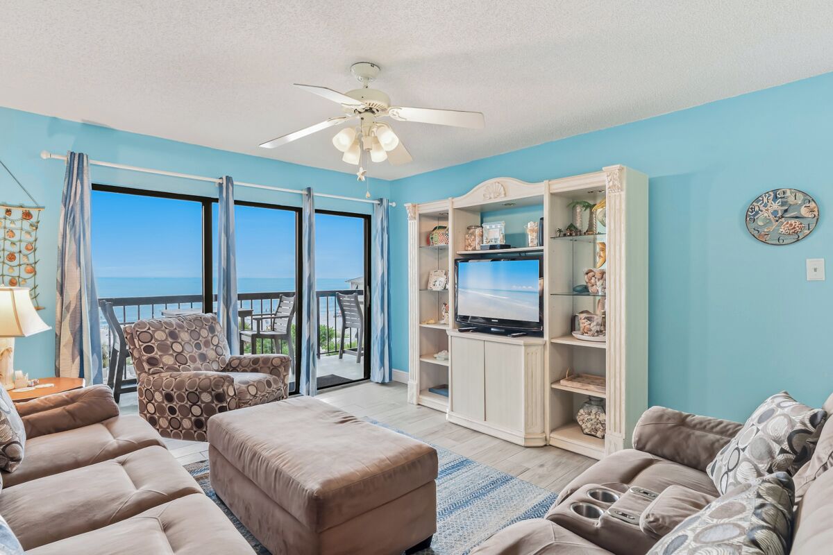 A living room with a large TV, comfortable seating, a ceiling fan, and a view of the outdoors.