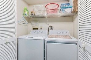 A laundry room with two stacked washers and dryers, a shelf with laundry baskets, detergent, and a light bulb box. - Top Floor Gulf Front • Pool • Sand Castle North 503 (Indian Rocks Beach)