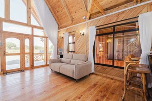 A wooden interior with a sofa and a chair, large windows, and a view of the outdoors.