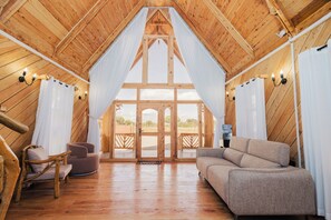 A wooden interior with a high ceiling, a sofa, a chair, and a view through a large window.