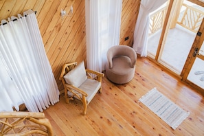 A room with wooden flooring, a round chair, a small sofa, and a large window with curtains.