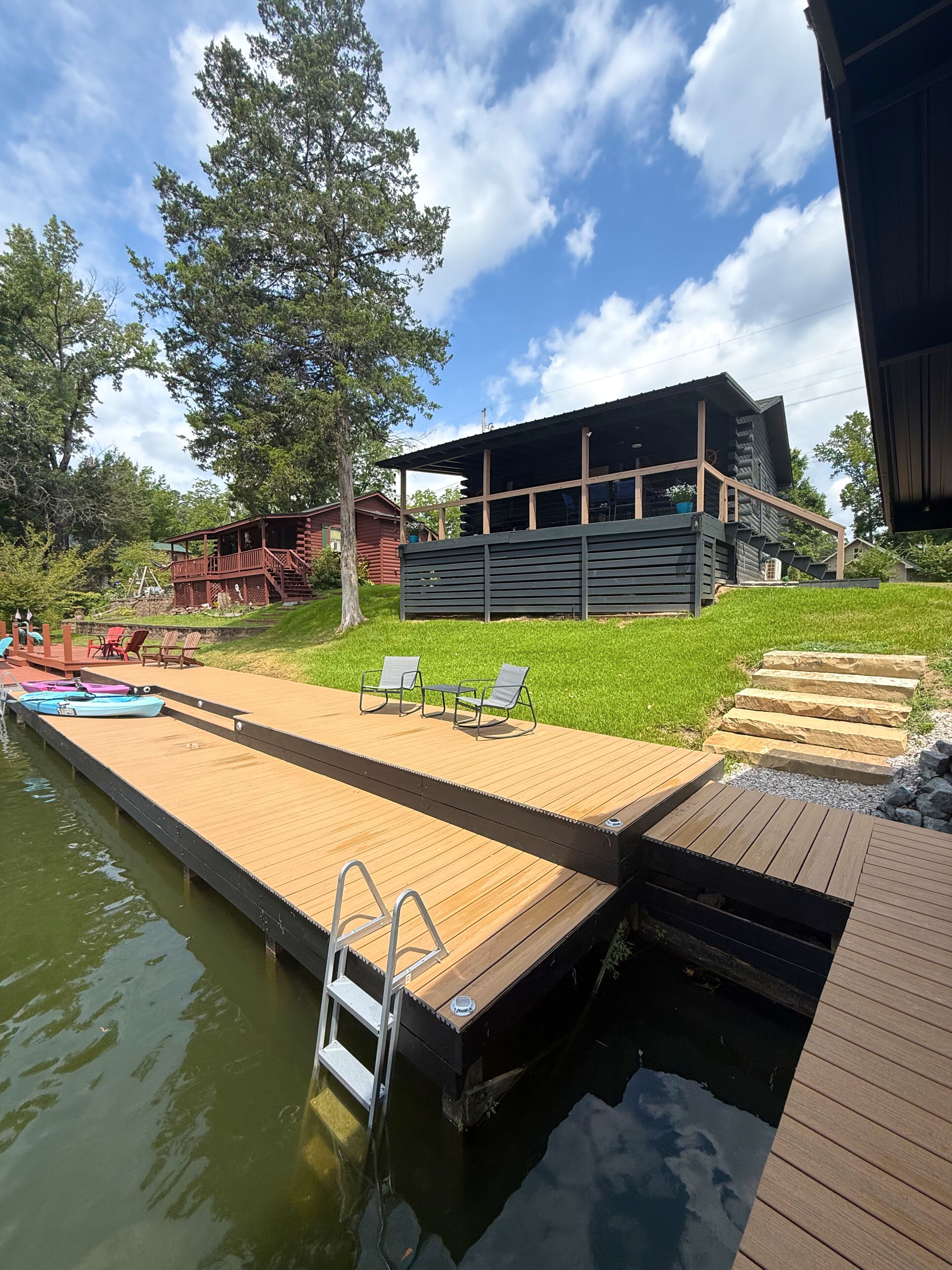 Een houten steiger die uitloopt in een wateroppervlak, met een modern gebouw en een boom op de achtergrond.