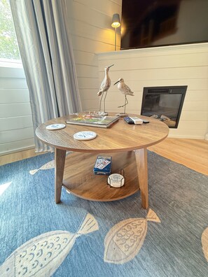 Ein runder Holztisch mit einem blau gemusterten Teppich, zwei Vogel-Skulpturen, einem Gesellschaftsspiel und einem kleinen Tischchen mit einem Zitat.