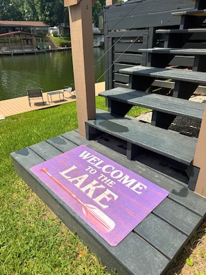 Eine Holzterrasse mit einer violetten Fußmatte zum See, eine Bank und im Hintergrund eine Seeblick mit Gebäuden.