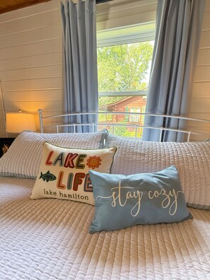 Ein Bett mit zwei Zierkissen, ein Fenster mit Vorhängen und eine Lampe auf einem Nachttisch.
