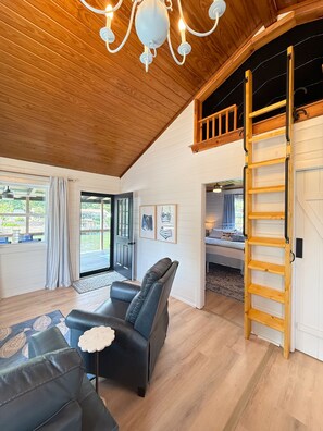 A spacious room with a wooden ceiling, a black leather sofa, a wooden ladder, and a view of a bedroom with a bed and a window. - Cozy Cabin on Lake Hamilton (Mountain Pine)