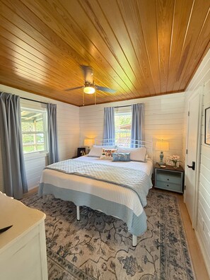 Ein Schlafzimmer mit einem großen Bett, einem Deckenventilator, einem Nachttisch mit Lampe und einem Fenster mit Vorhängen.
