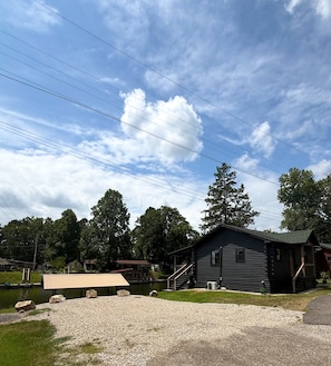 A rustic cabin with a gravel driveway and a covered area, surrounded by trees and a body of water. - Cozy Cabin on Lake Hamilton (Mountain Pine)