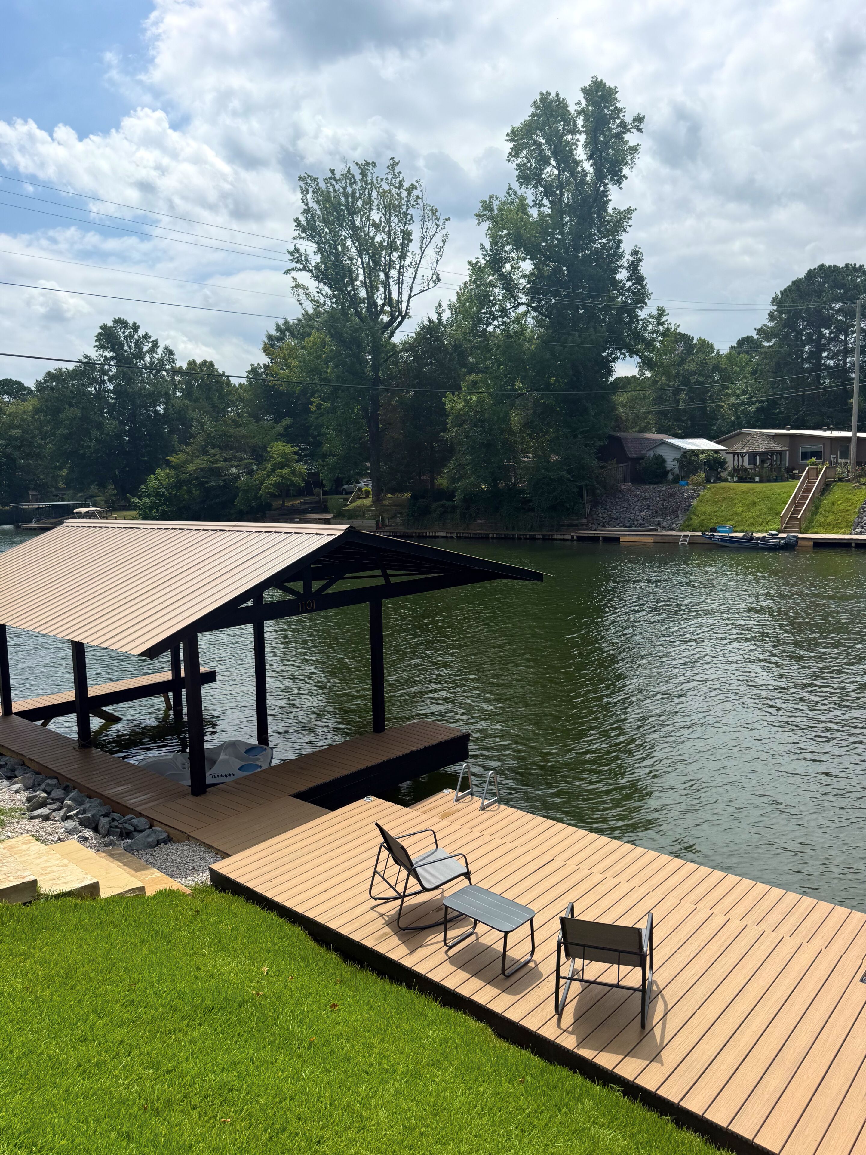 A wooden dock with a covered seating area, a boat, and a grassy bank.