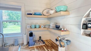 A kitchen with a wooden ceiling, white walls, and a window. There are shelves with dishes and kitchen utensils.