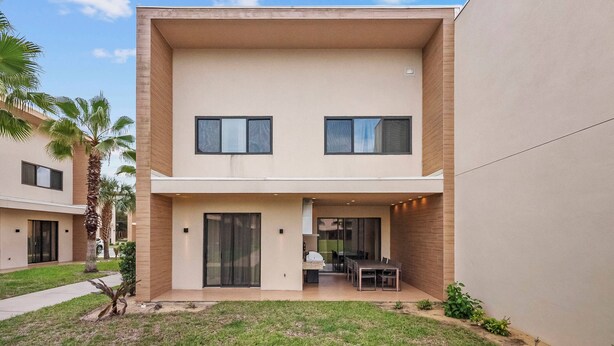 Ein modernes, zweistöckiges Gebäude mit flachem Dach, großen Fenstern und einer überdachten Terrasse mit Gartenmöbeln.