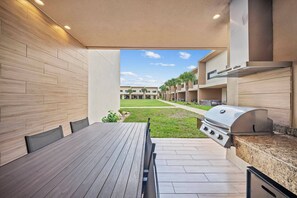 Een terras buiten met een houten tafel en stoelen, een barbecue en een uitzicht op een gebouwencomplex in de verte.