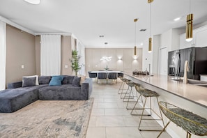 Een moderne woonkamer met een hoekbank, een eethoek met een tafel en stoelen, en een keuken met een koelkast en barstoelen.