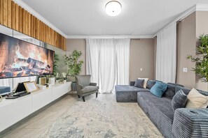 Een moderne woonkamer met een flatscreen televisie, een grijze bank, een houten accentmuur en een groot raam met gordijnen.
